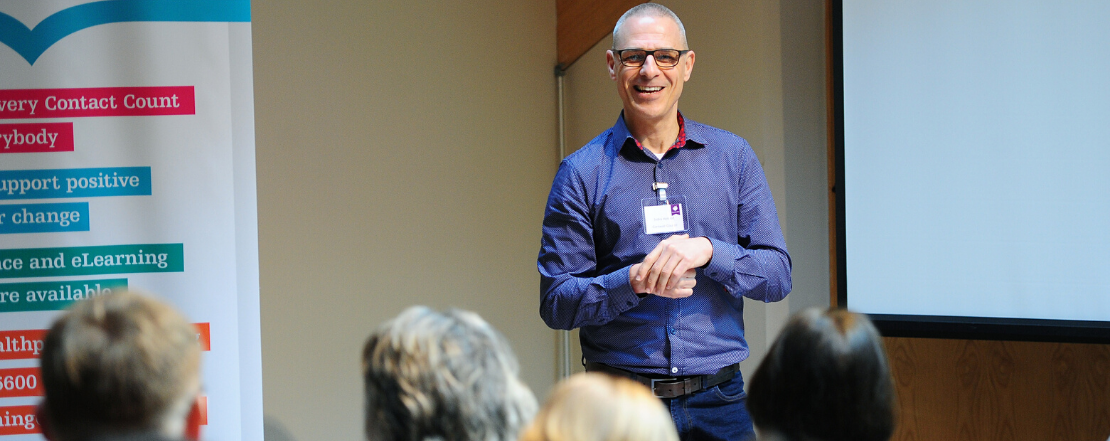 man stood at the front of a room of people giving a training presentation